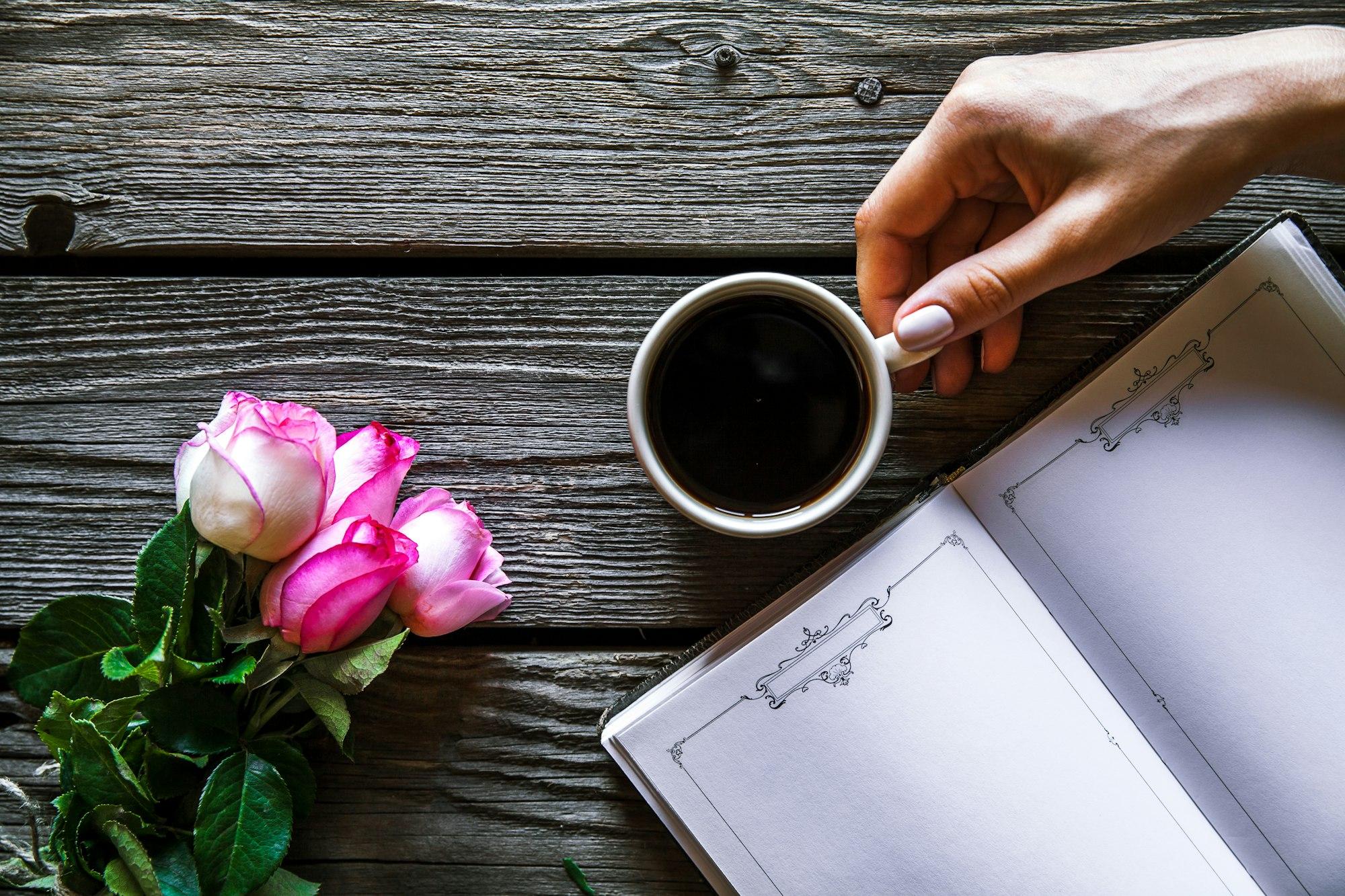 Female hand with a cup of coffee, book and flowers on wooden background. Flowers, break, work